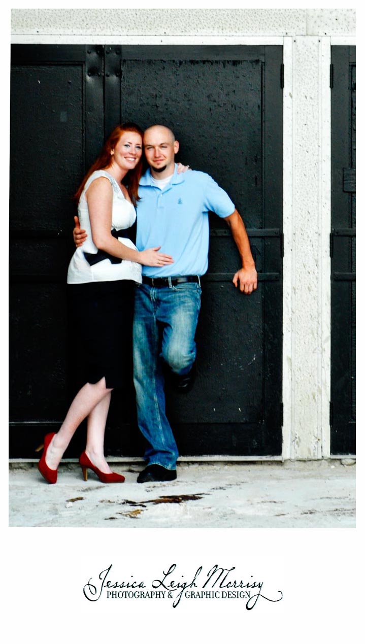 Couple stands closely in front of black double doors, smiling warmly as the woman leans into the man, her red heels adding a pop of color.