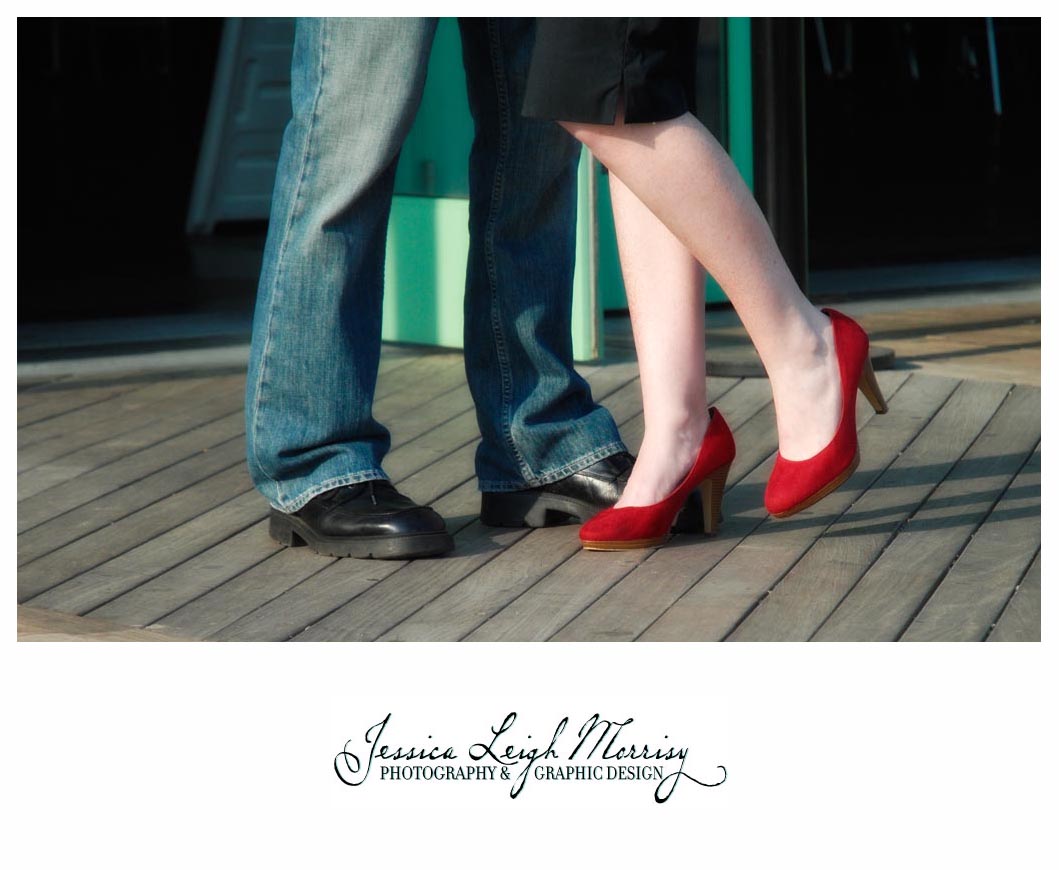 Close-up of couple's feet on a boardwalk—her red heels and his black shoes with jeans—capturing a playful, romantic moment.