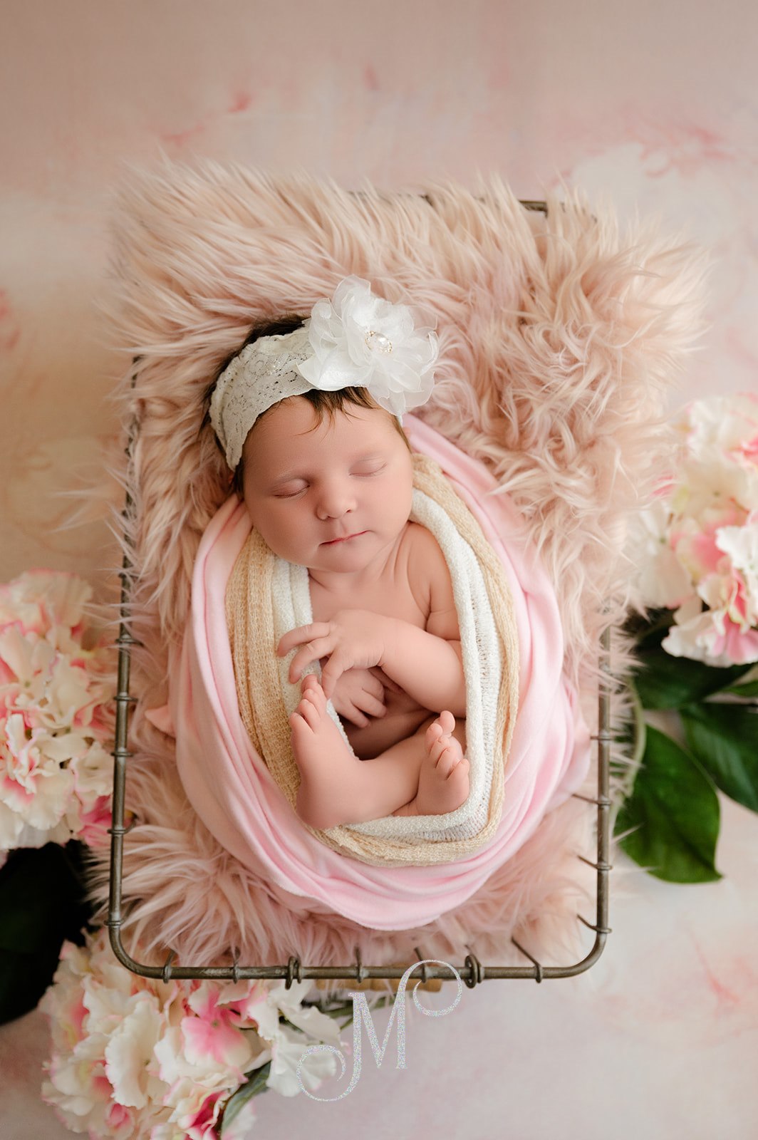 Sleeping newborn baby girl wrapped in soft pink layers, resting on a fluffy blanket with delicate flowers and a floral headband in a timeless studio portrait.